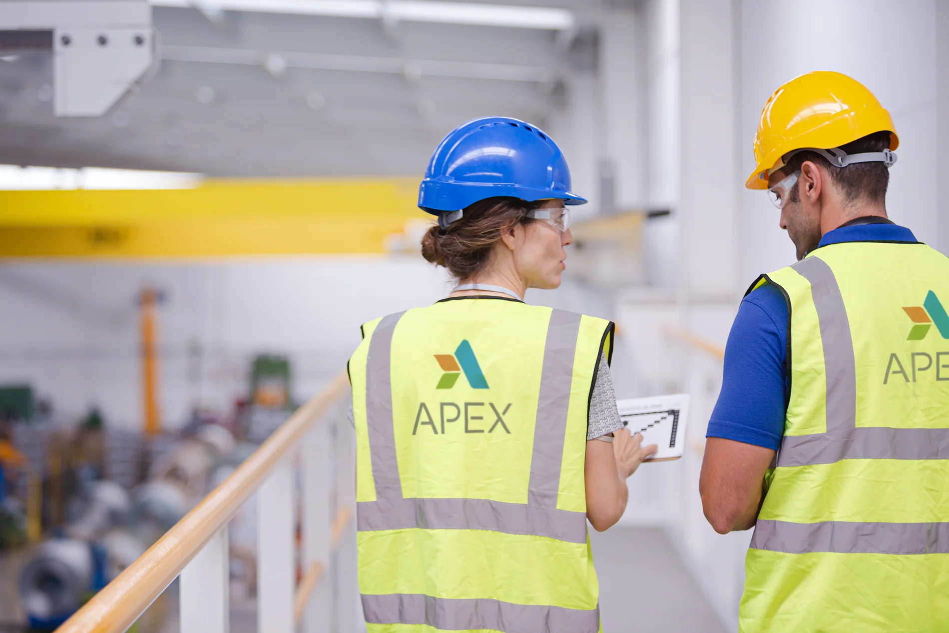 Apex workers wearing hardhats, protective eyewear, and safety vests discussing health and safety commitment goals inside a warehouse.