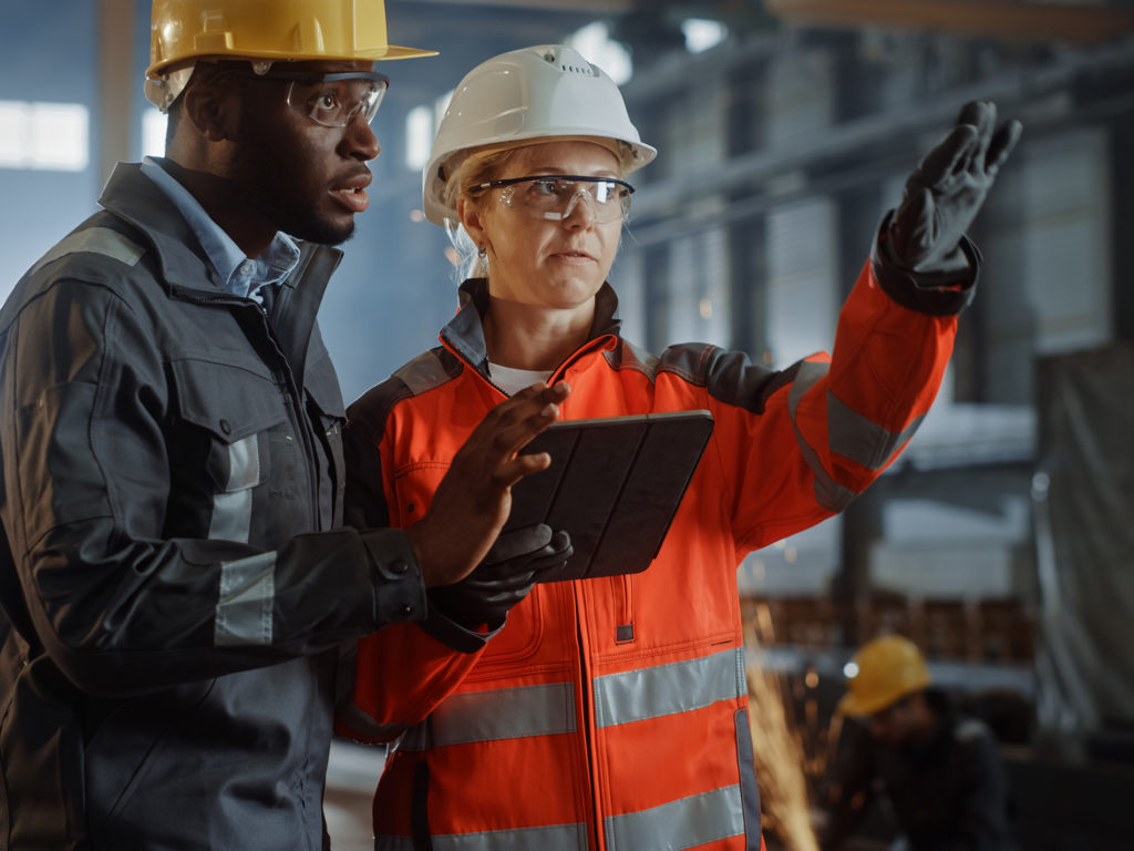 Two Heavy Industry Engineers Stand in Steel Metal Manufacturing Two Heavy Industry Engineers Stand in Steel Metal Manufacturing Factory, Use Digital Tablet Computer and Have a Discussion. Black African American Industrial Specialist Talk to Female Technician.