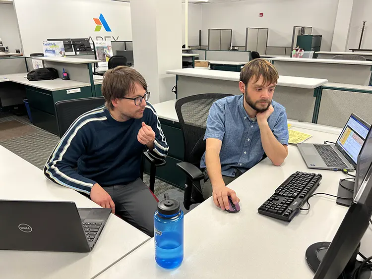Two Apex Companies team members in an office having a discussion at a desk in front of a computer